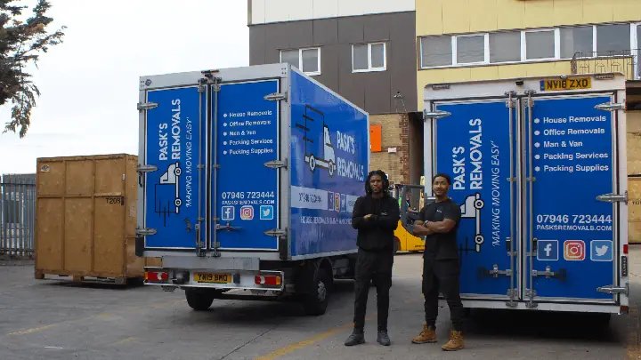 Two Pask's Removals London movers standing between branded box vans in a yard with wooden storage crates