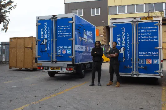 Two Pask's Removals London movers standing between branded box vans in a yard with wooden storage crates