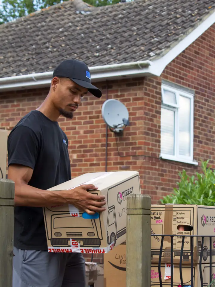 Pask's Removals London mover carrying a cardboard box past a brick house