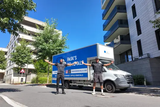 Two Pask's Removals London movers with arms outstretched beside the van among modern apartment blocks
