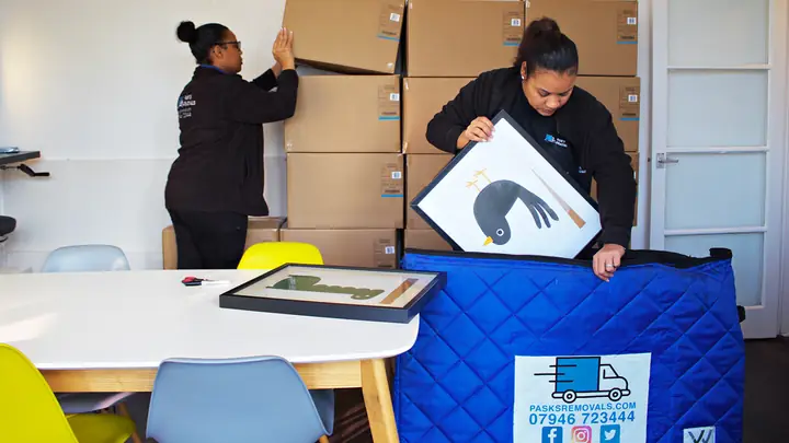 Two Pask's Removals London packers wrapping framed pictures in blue blankets beside stacked boxes