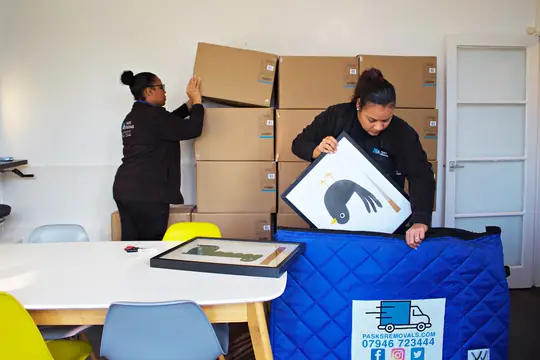 Two Pask's Removals London packers wrapping framed pictures in blue blankets beside stacked boxes